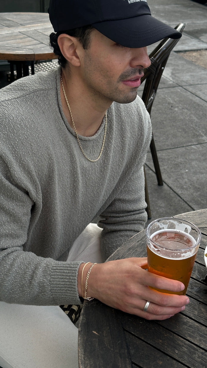 A man wearing a baseball cap, grey sweater, and assorted silver bracelets holding a beer on a table