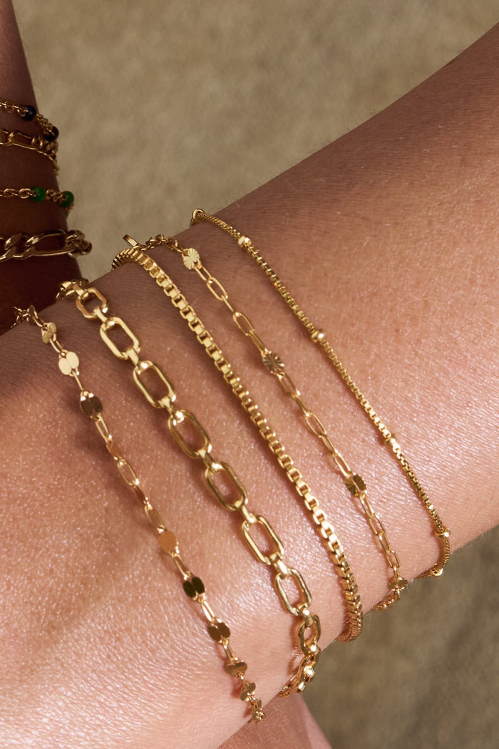 Close up of a wrist with dainty gold chain bracelets on a tan background