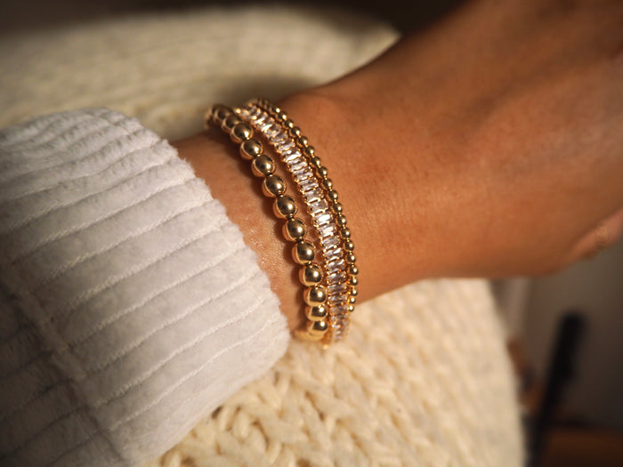 Close up of a wrist in a white sweater with gold and stone bracelets resting on a white knit blanket