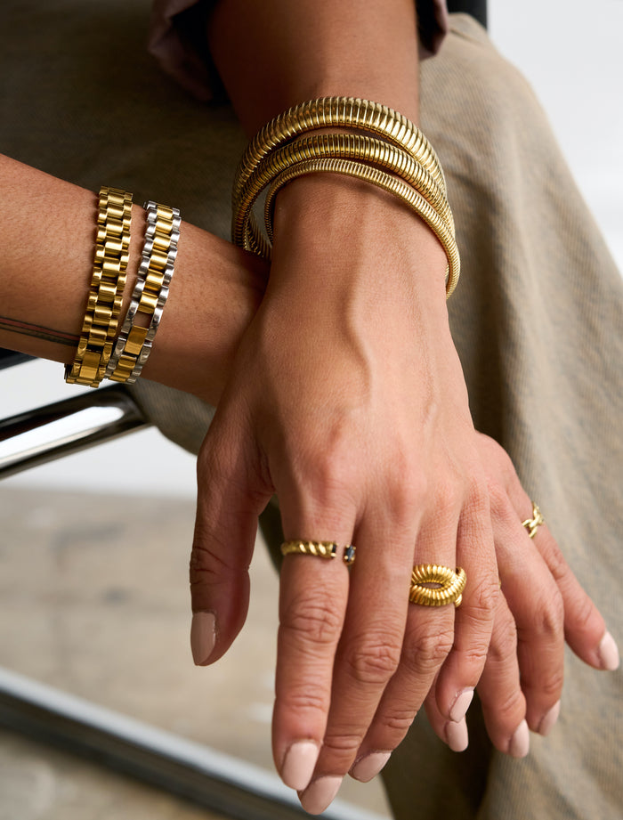 Two wrists with gold rings and bracelets resting on the arm of a chair