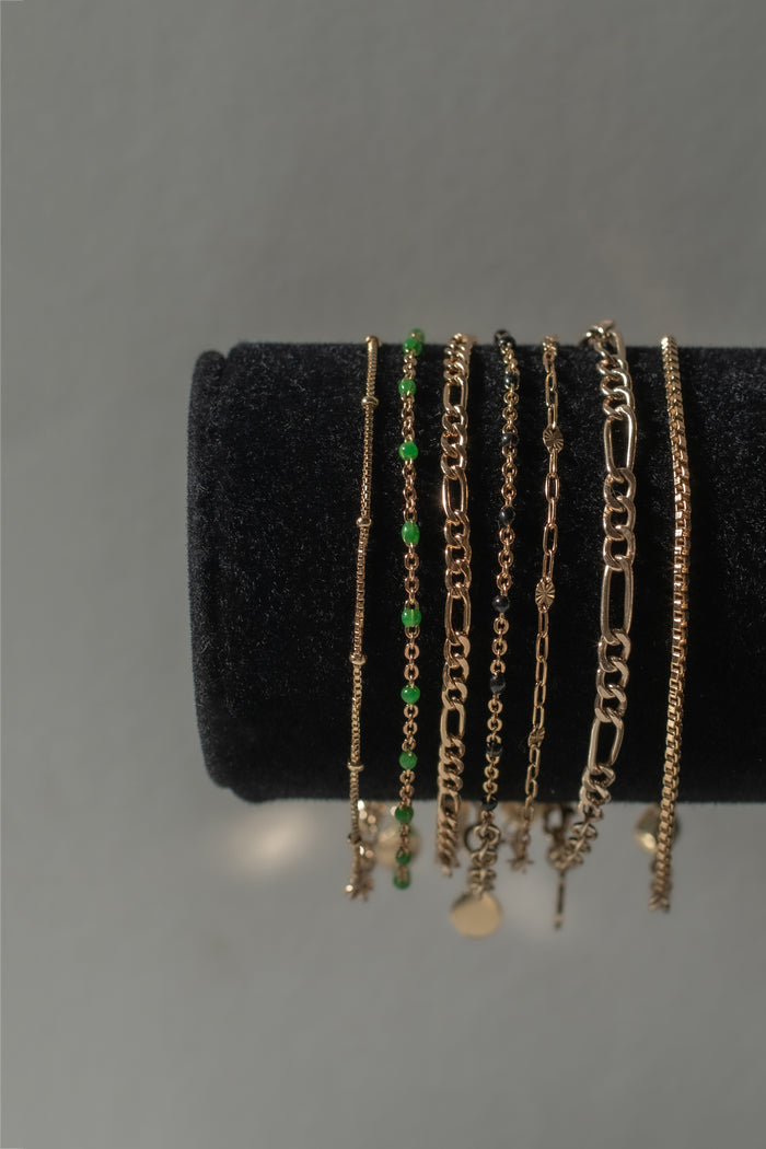 Close up of dainty gold chain bracelets resting on a black cuff on a grey background