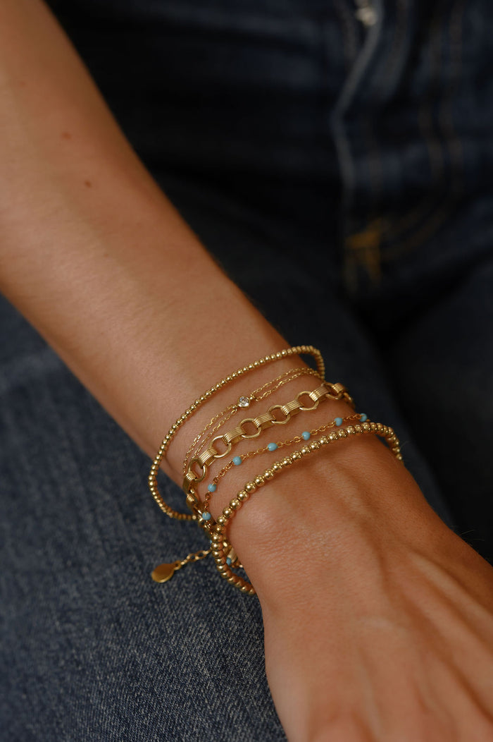Close up of a wrist with assorted gold bracelets with stone accents resting on a pair of jeans