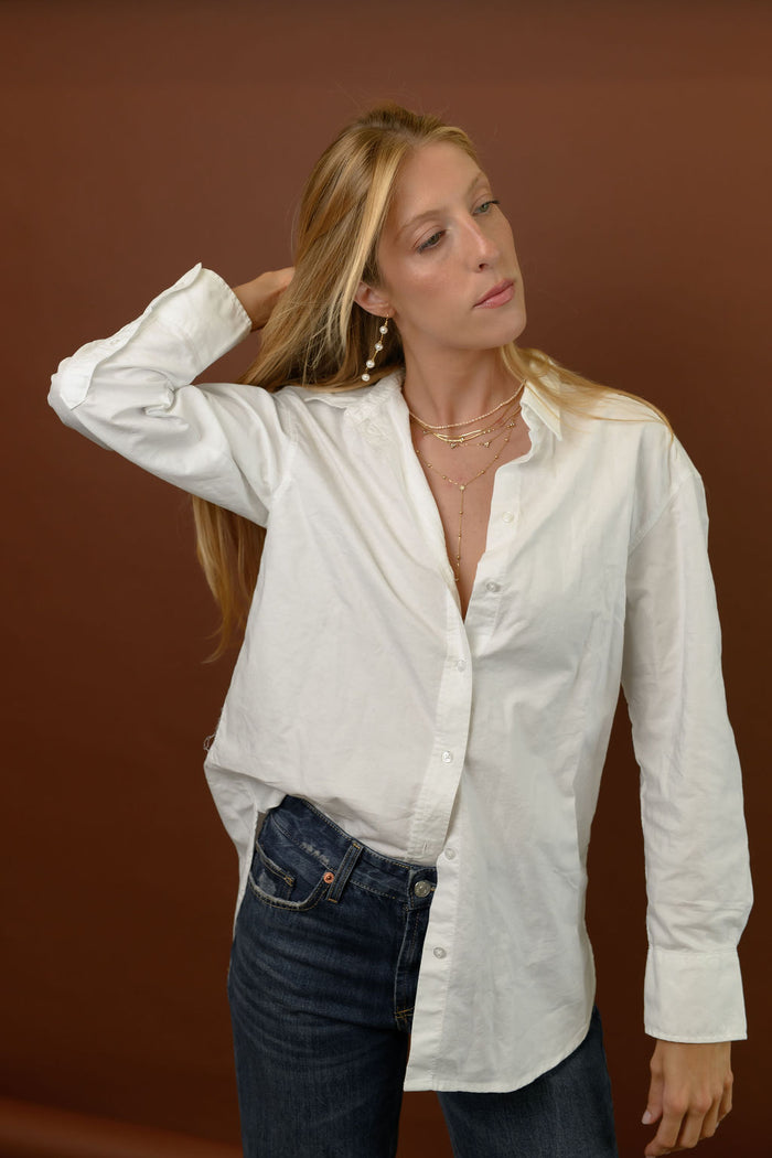 A woman wearing gold jewelry and a white button down with black jeans on a brown background