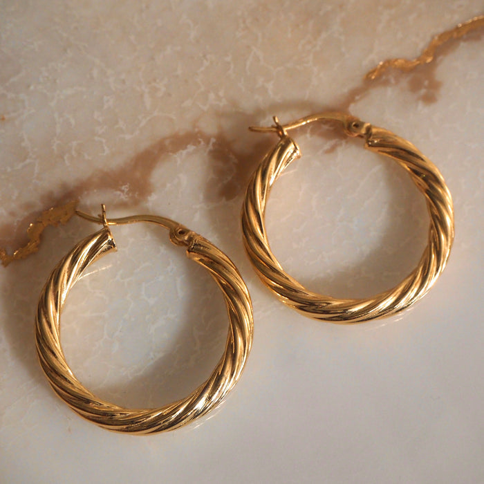 Close up of twisted gold hoop earrings on a neutral stone background