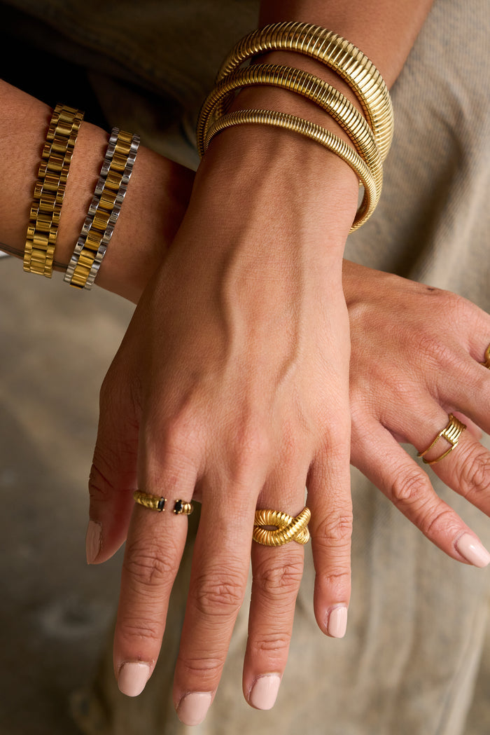 Two wrists with gold rings and bracelets on a khaki background