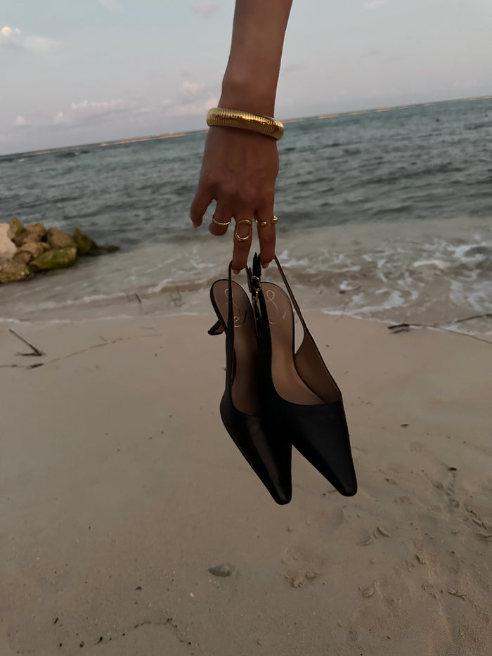 A hand with gold bracelets and rings carrying a pair of black heels on the beach