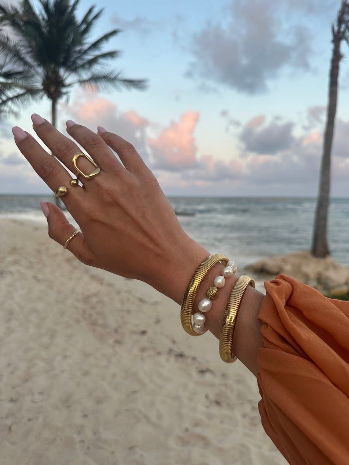 A wrist in a orange blouse with gold rings and bracelets on the beach