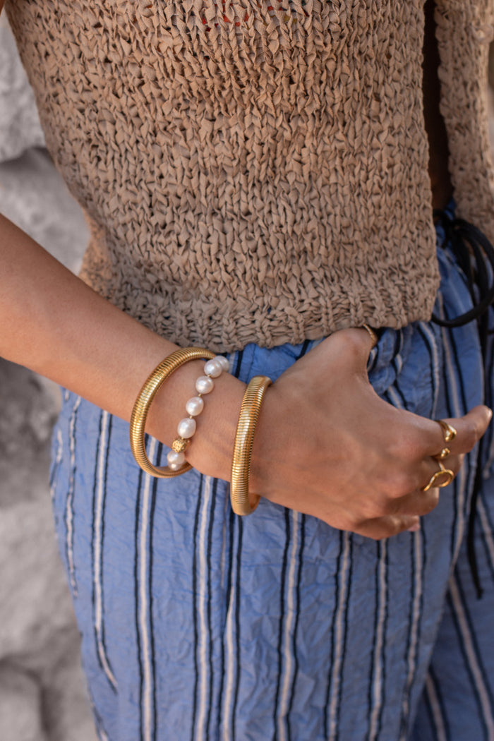 A person in a vest and striped linen pants wearing gold and pearl bracelets and rings