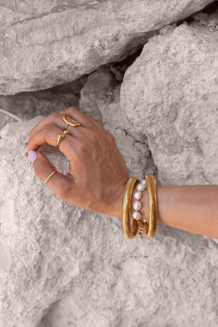 A hand with gold and pearl rings and bracelets resting on a rock