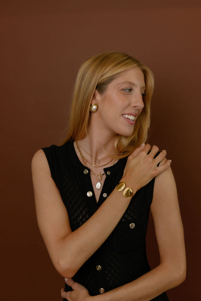 A woman wearing gold accent earrings, necklaces, and bracelets on a brown background
