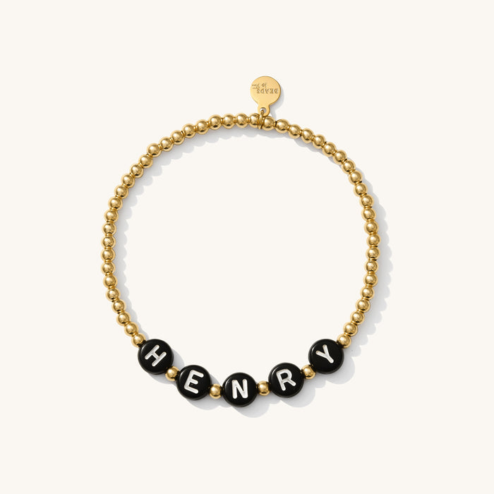Close up of a gold bead bracelet with white letter beads reading "HENRY"