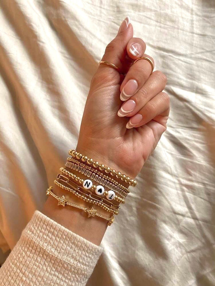 A wrist with assorted gold and stone bracelets, including a custom bracelet that says "T<3N"