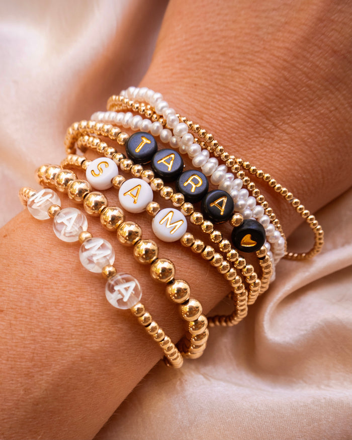 A wrist with gold bead bracelets, including personalized name bracelets on a tan cloth that read "TARA <3", "SAM", and "MAMA"