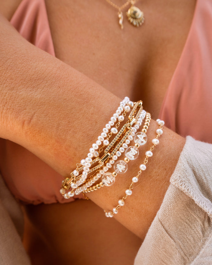 A person wearing a pink top, assorted gold and pearl bracelets, and a custom name bracelet that reads "MAMA"