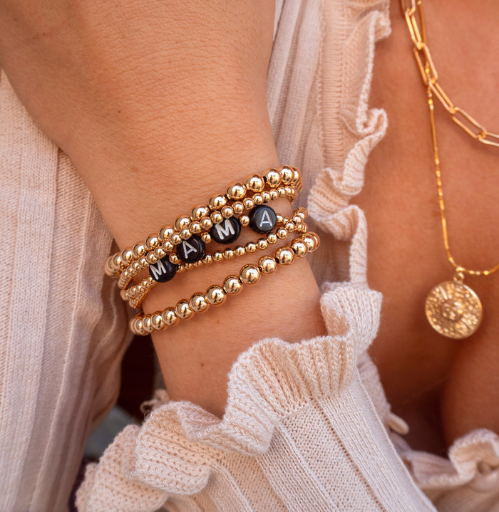 Close up of a woman wearing a tan blouse and gold jewelry, including necklaces, bracelets, and a custom bracelet that says "MAMA"