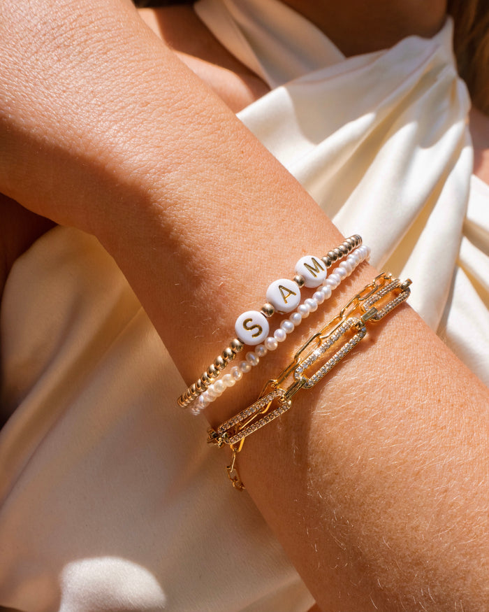 Close up of a person wearing a white halter top and gold bracelets, including a custom bead bracelet that says "SAM"