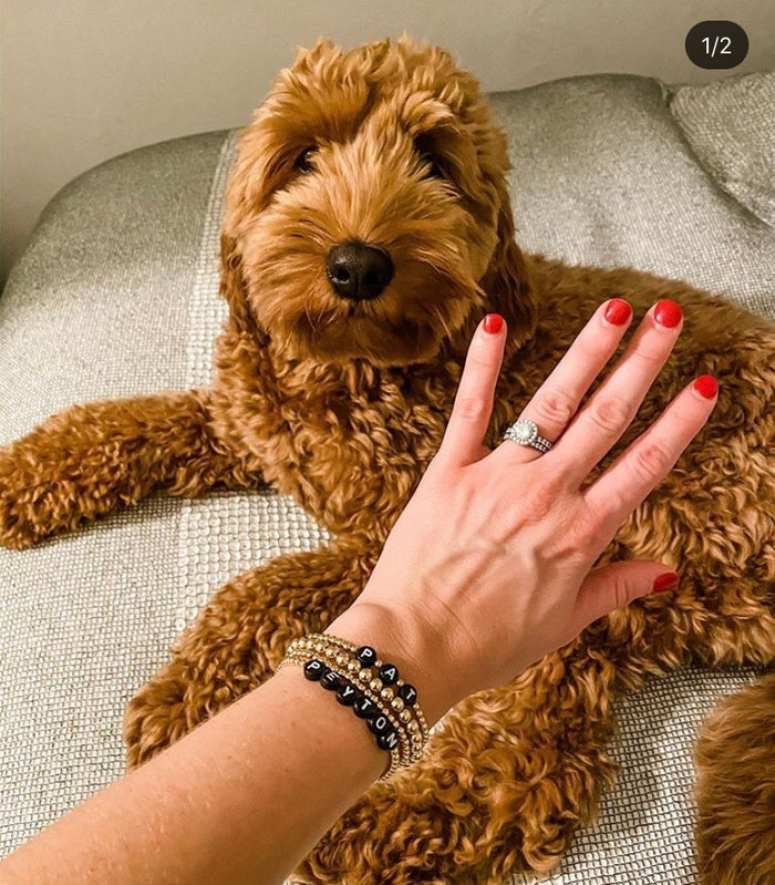 A person's wrist with custom name bracelets that say "PAT" and "PEYTON" with a dog in the background