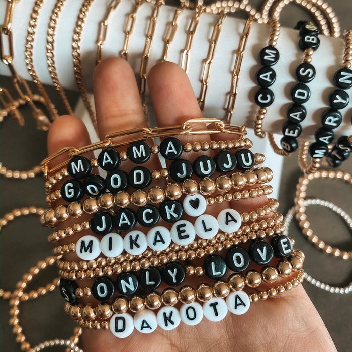 A hand holding assorted custom gold bead bracelets, with more bead bracelets in the background