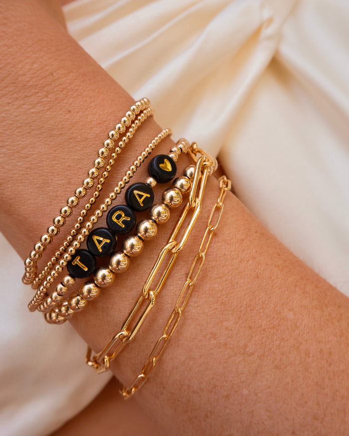 Close up of a wrist with assorted gold bracelets, including a gold bracelet with black beads that reads "TARA <3"