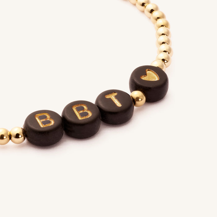 Close up of a gold bracelet with black beads that say "BBT <3"