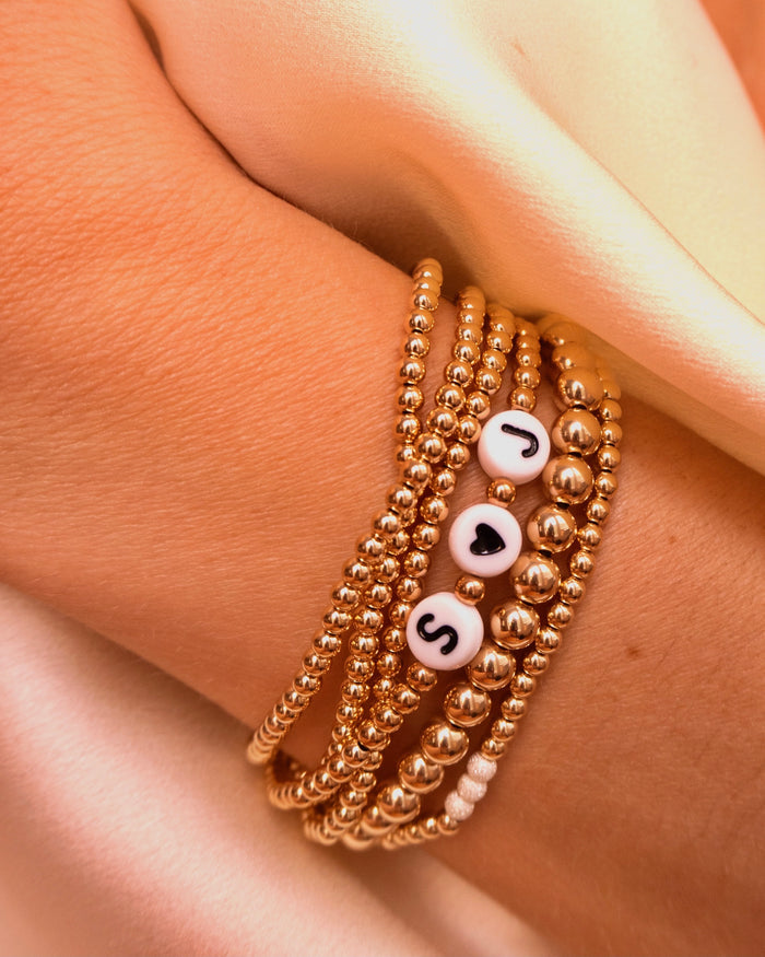 Close up of a wrist with gold bead bracelets, including a custom bead bracelet that reads "S<3J", on a white background