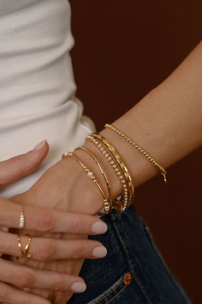 Close up of a wrist with multiple gold and pearl bracelets on a brown background