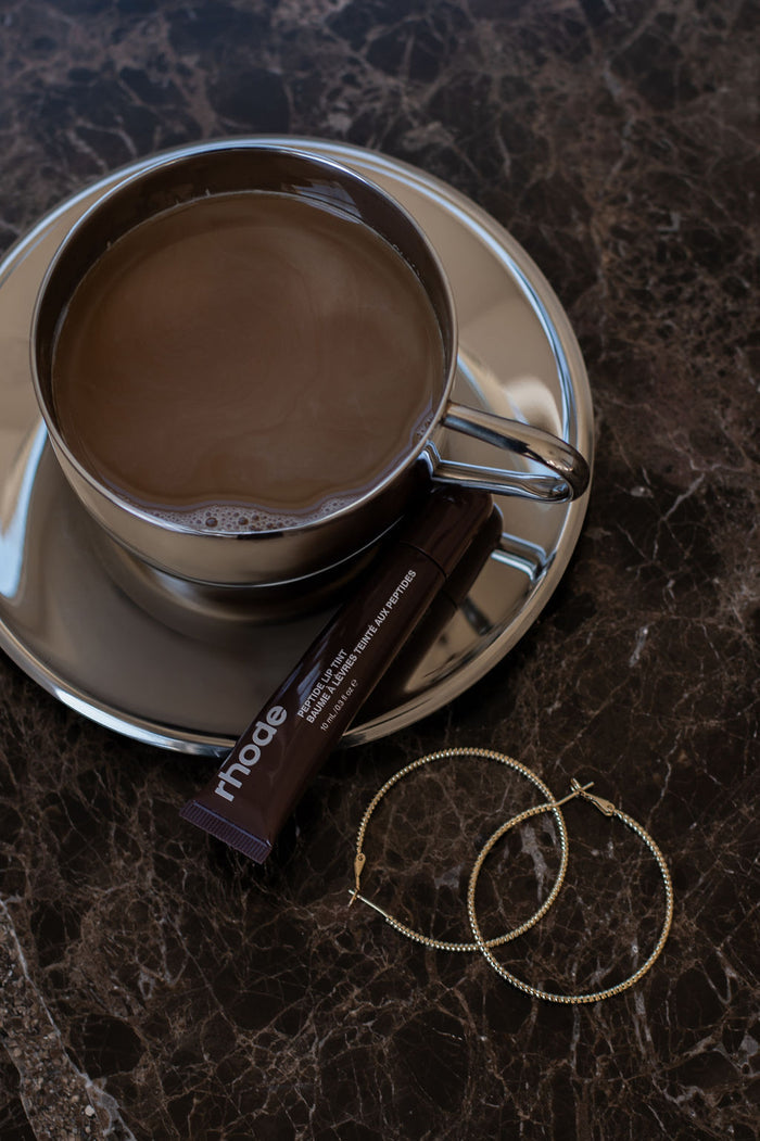 A coffee cup with coaster, lip gloss, and hoop earrings on a marble table