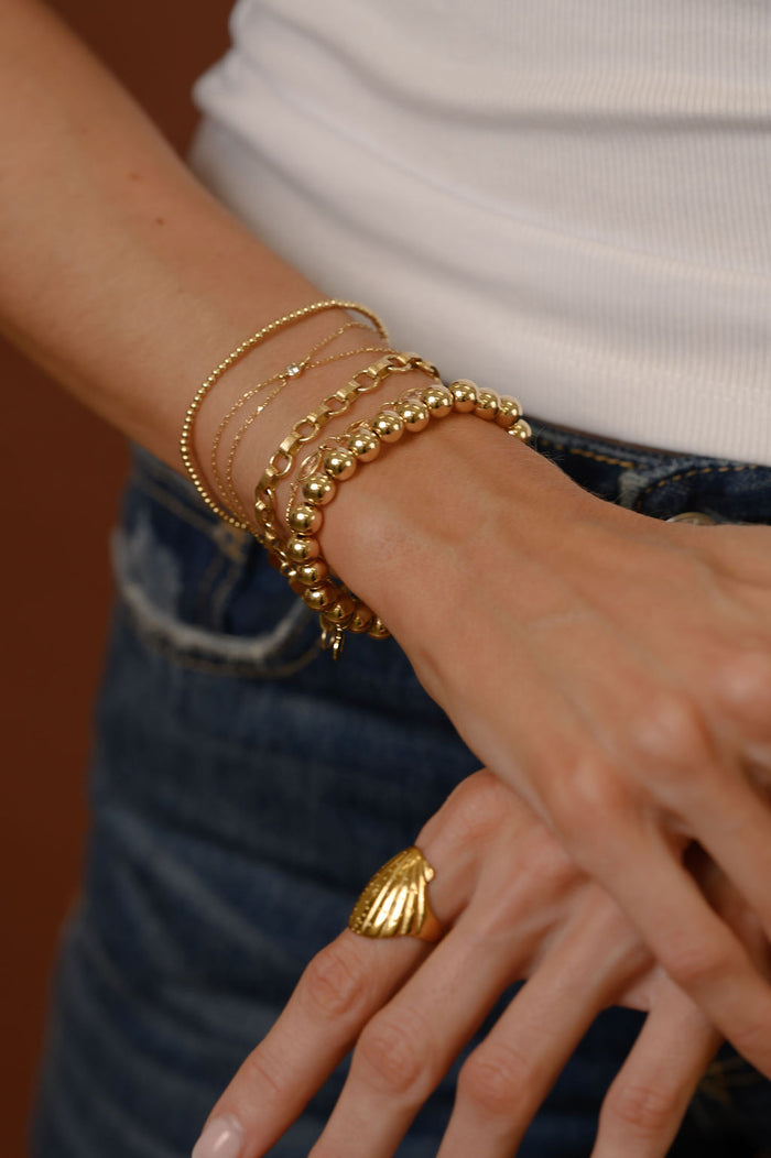 Close up of a person wearing gold bracelets, a white tank top, and blue jeans against a brown background
