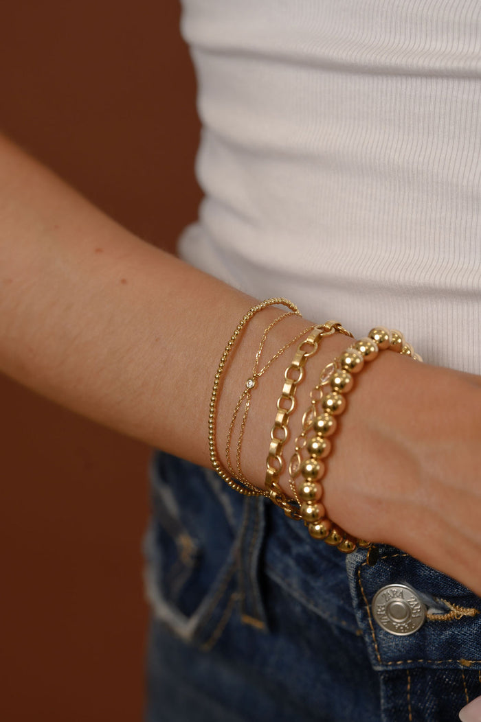 Close up of a person wearing gold bracelets, a white tank top, and blue jeans against a brown background
