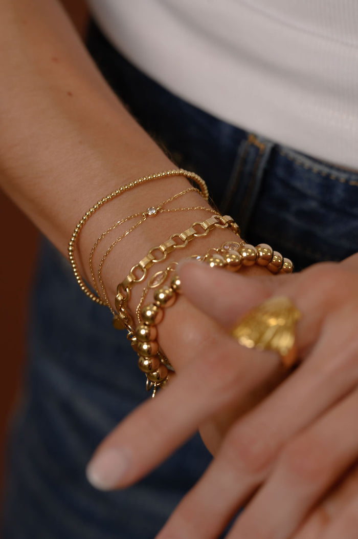 Close up of a person wearing gold bracelets, a white tank top, and blue jeans against a brown background