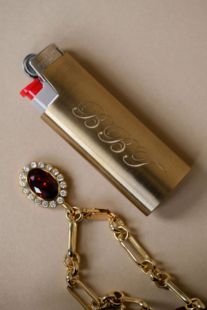 Gold lighter with 'BBJ' branding and a decorative keychain on a beige background