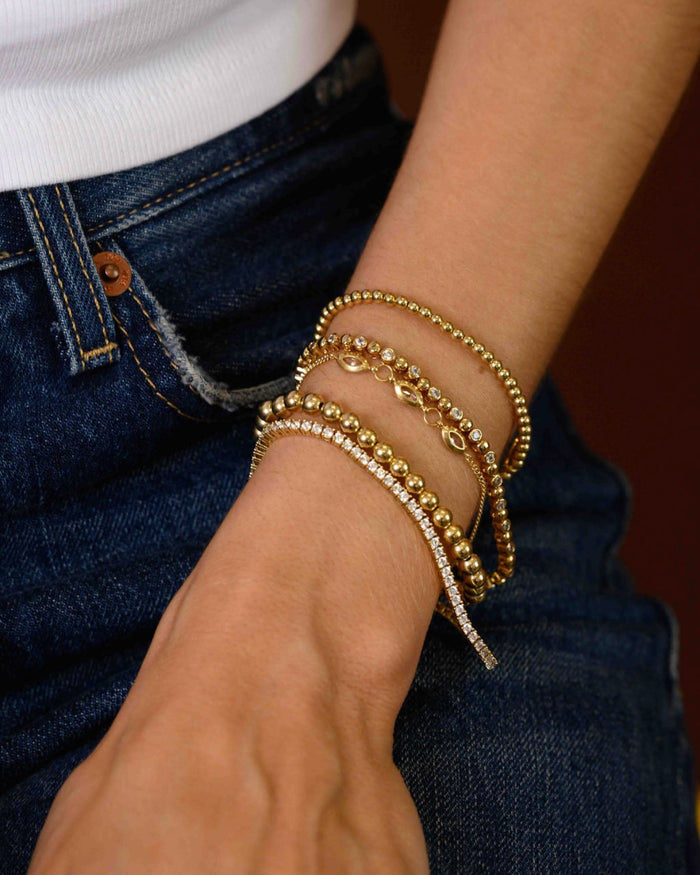 Close up of a person wearing a white shirt, blue jeans, and assorted gold and stone bracelets against a brown background