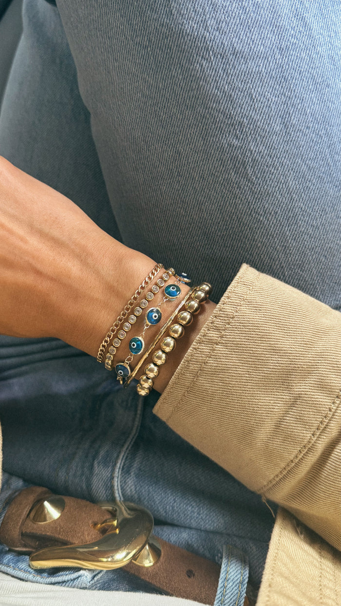 Close up of a woman's wrist with a tan jacket and assorted gold bracelets resting on a pair of jeans