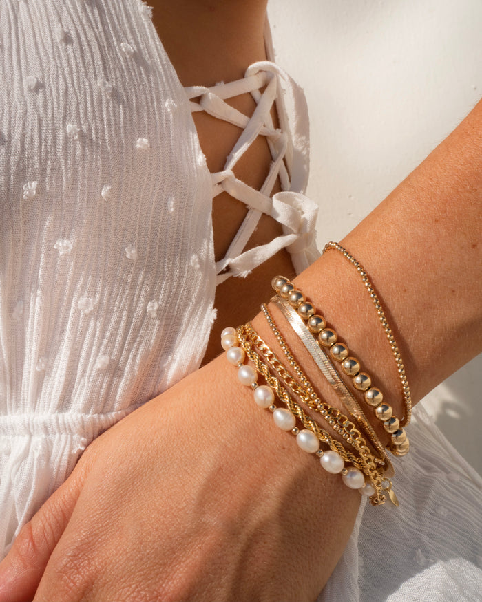 Close up of a person wearing a white top, holding up their wrist with assorted gold and pearl bracelets