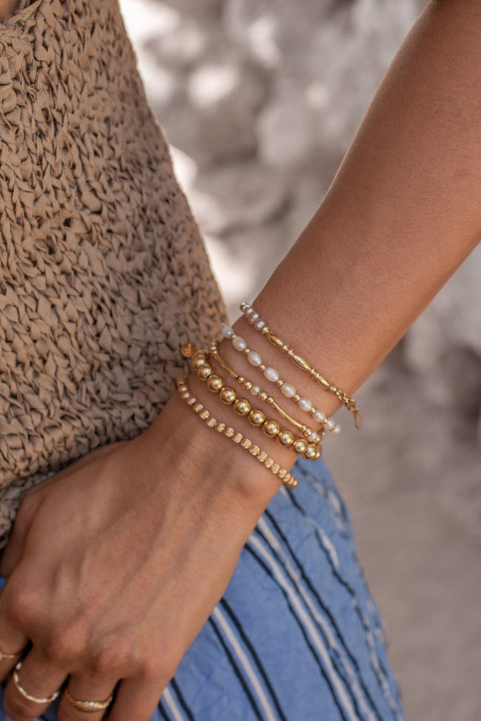 A person wearing a brown top, blue pants, and assorted gold bracelets and rings on a tan background