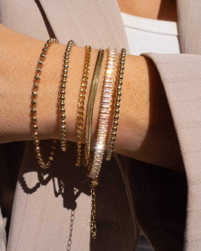 Close up of a person's chest and wrist wearing a tan blazer, white top, and assorted gold and stone bracelets