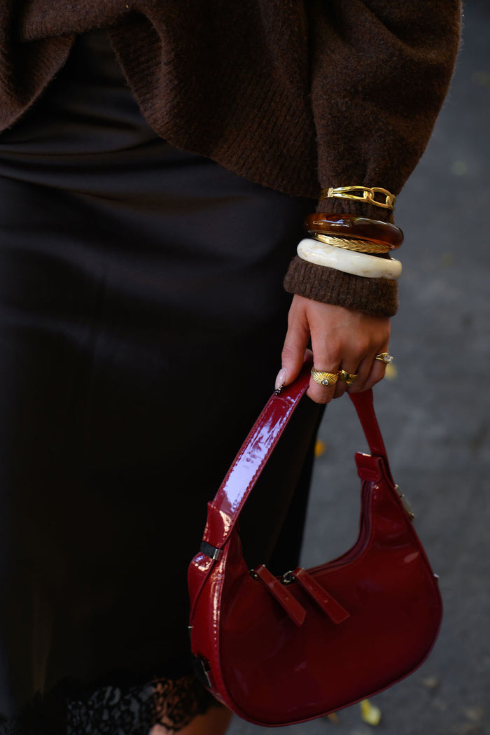 A person in a brown sweater and black skirt with assorted gold and stone jewelry, holding a red purse