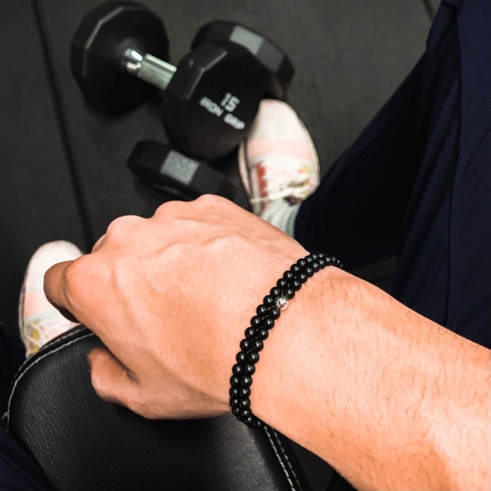 A person wearing black pants, white sneakers, and black bracelets sitting on a gym bench, with a set of weights at his feet