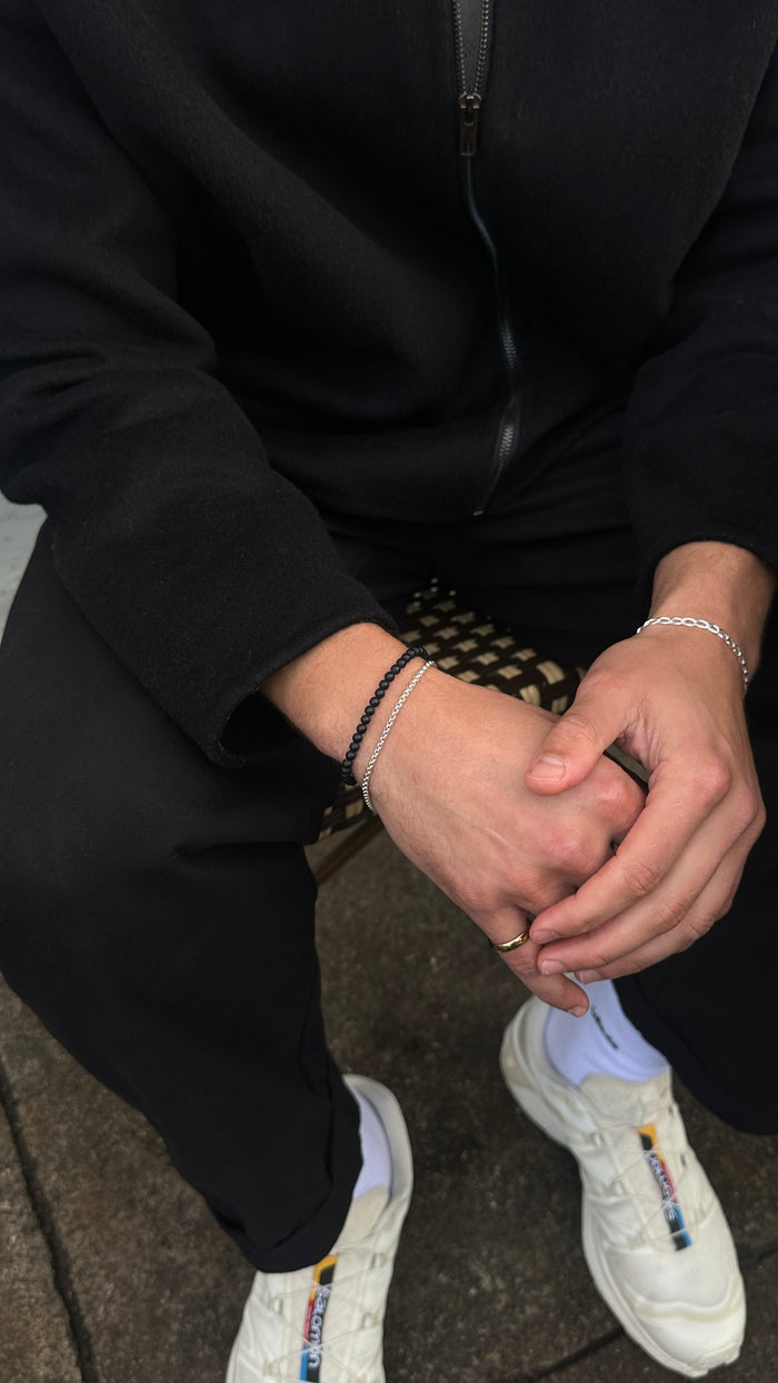 A person wearing an all-black outfit and white shoes sitting on a chair, accessorized with black and silver jewelry