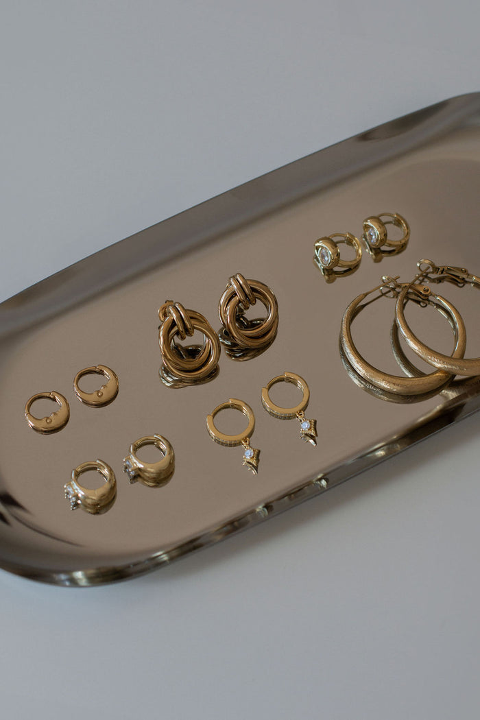 Six pairs of assorted gold earrings on a silver tray set on a grey background.