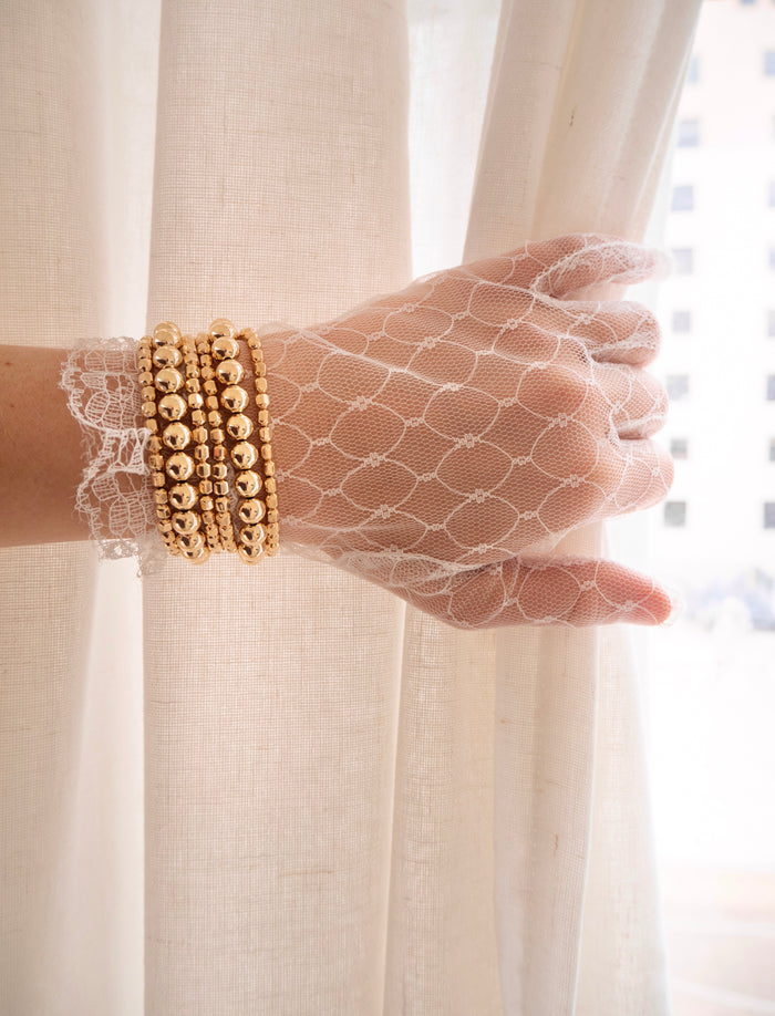 A person wearing a lace glove and gold bead bracelets holding a white curtain