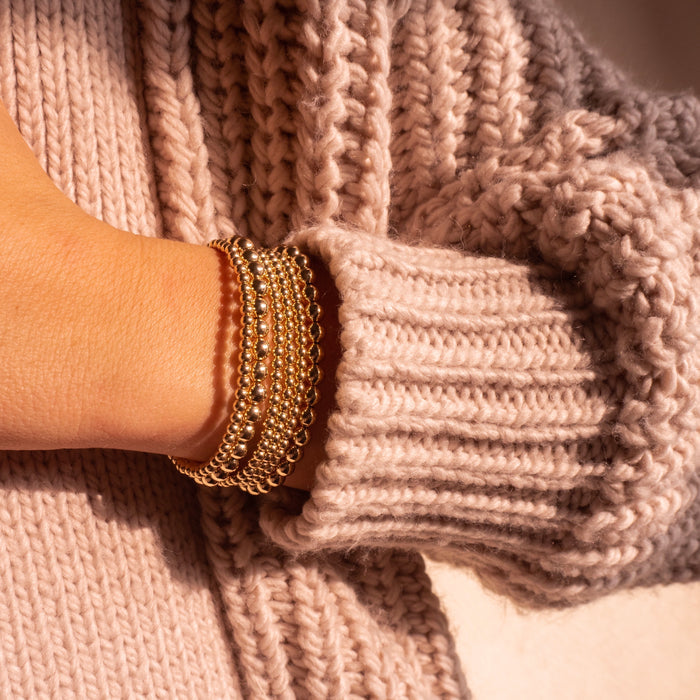 Close up of a person's torso and arm wearing a pink crochet sweater and gold bead bracelets