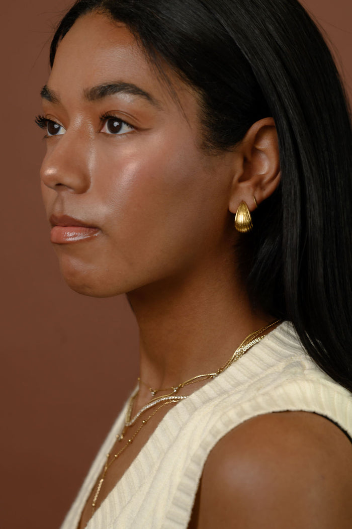 A woman wearing gold earrings and necklaces against a brown background