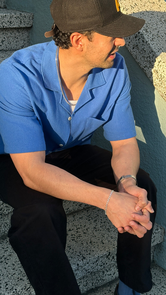 A person sitting on a staircase wearing a black baseball cap, blue shirt, black pants, and silver bracelets