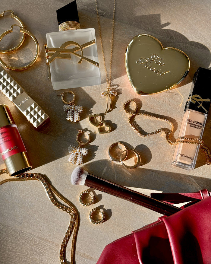 A flatlay shot of makeup, perfume, and gold jewelry on a tan background