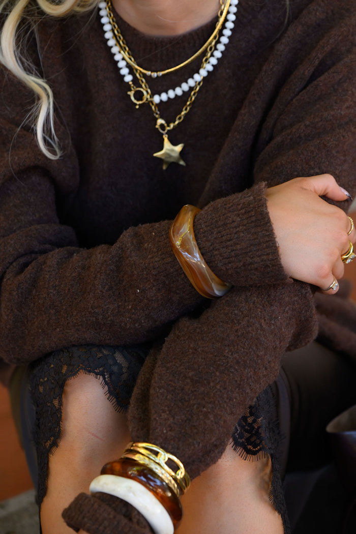 Close up of a person in a brown sweater with gold and resin rings, bracelets, and necklaces