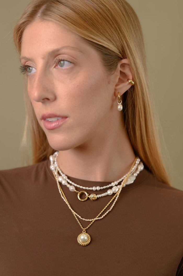 A woman wearing gold and pearl earrings and necklaces in a brown shirt against a green background
