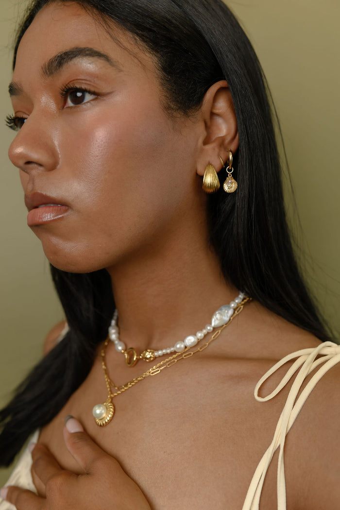 A woman wearing gold and pearl earrings and necklaces against a green background