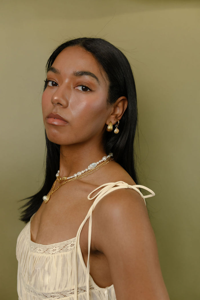A woman wearing gold and pearl earrings and necklaces in a white blouse against a green background