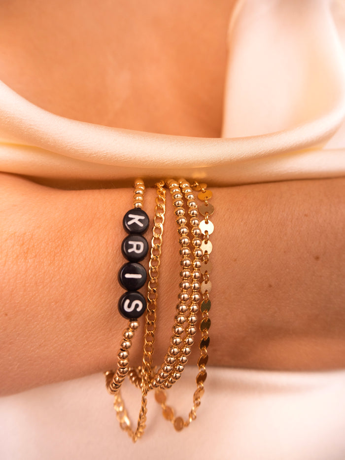 Close up of a person's wrist with gold bracelets, including a custom bracelet reading "KRIS", up against a white background